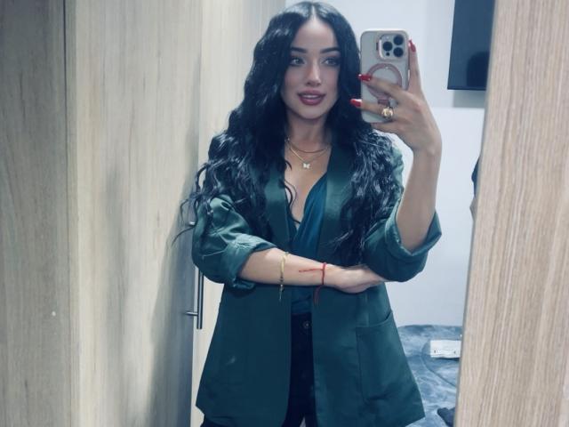LillyTaylor with long black wavy hair, green shirt, black dress, red nails, holding phone, mirror selfie, necklace, bracelet, and ring, standing in wooden room.