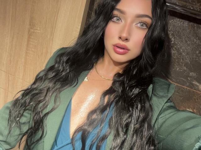 LillyTaylor with long black wavy hair, green jacket, blue top, shiny skin, pink lips, and green eyes, wearing a gold necklace, looking directly at the camera.