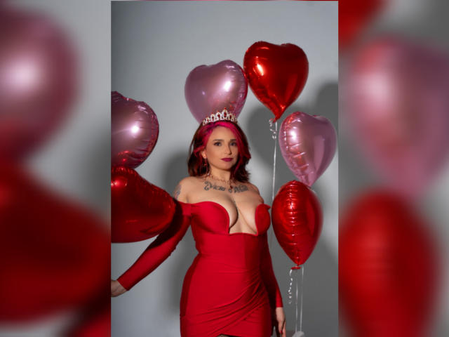Alexandrapsique in a red off-shoulder dress, holding red and pink heart balloons, wearing a silver tiara, with tattoos on her chest, and red lipstick.