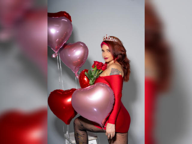 Alexandrapsique with red dress, tiara, holding pink and red heart balloons, black fishnets, and roses, showing her tits and a tattoo on her right arm.