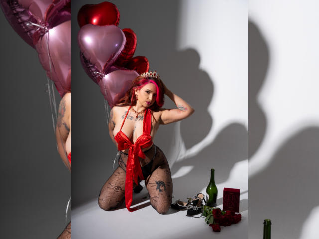 Alexandrapsique with pink hair, red lingerie, and a tiara, kneeling, holding heart balloons, tattoos, fishnet stockings, and a bottle, with gifts and red box on the floor.