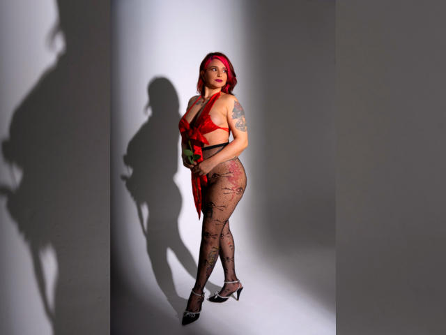 Alexandrapsique with red hair, wearing a red and green outfit, black lace stockings, and black heels, standing, showing her ass and tattoos, shadow on the wall.