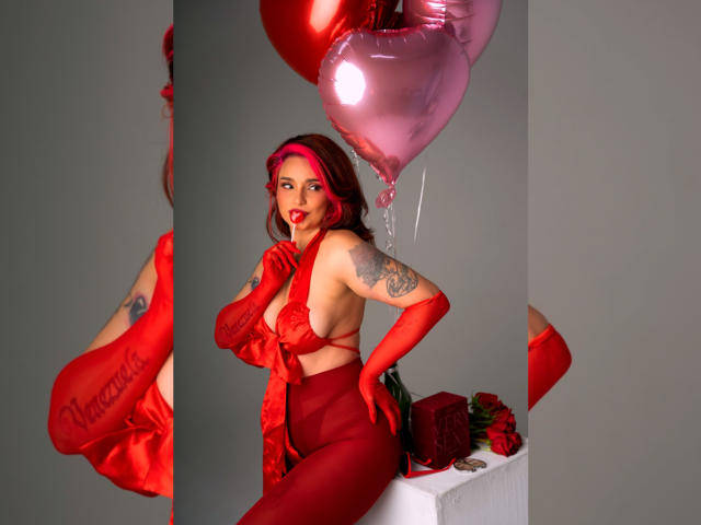 Alexandrapsique in red lingerie, gloves, and red rose headband, holding pink balloons, with tattoos on her arm, showing her tits and ass, next to roses on a white table.