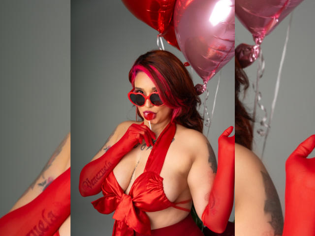 Alexandrapsique with red lips, red heart-shaped glasses, red satin bra exposing her big tits, red gloves, pink balloons, and tattoos, posing with finger to mouth, sexy and seductive.