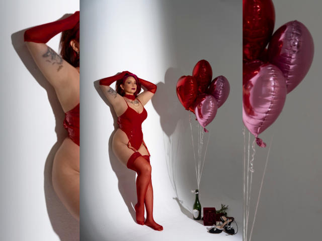 Alexandrapsique in red lingerie, garter, and stockings, with tattoos, standing and posing with hands on head, next to pink heart balloons, and wine bottle on white background.