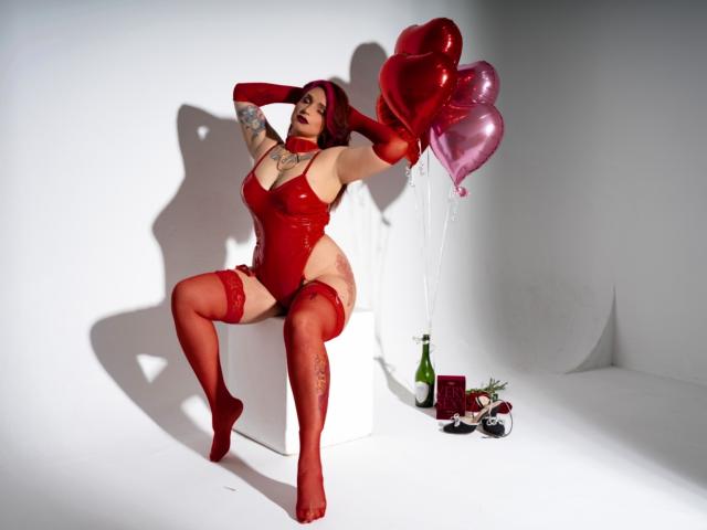 Alexandrapsique in red latex lingerie, holding pink heart balloons, sitting on white cube, arms raised, showing tattoos, red stockings, bottle, and decorations on floor. Boobs, ass, thigh tattoos.
