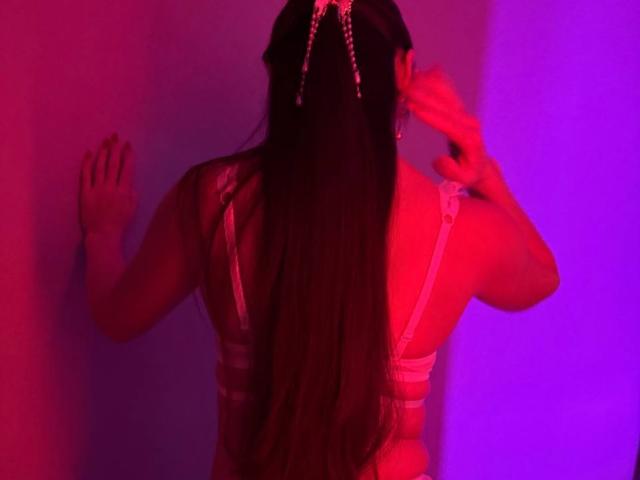 Rouzi with long black hair, back to camera, wearing a white lace bra, hands on a wall, illuminated by red and purple lights, showing her tits and ass.