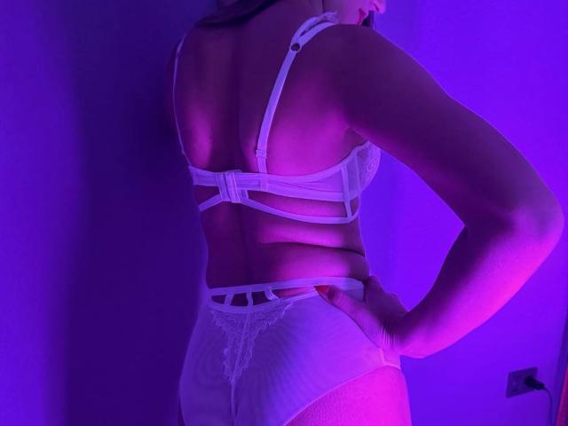 Rouzi's back is to the camera, wearing white lingerie with straps and lace, her ass slightly visible, under purple and pink lighting, hands on her hips.