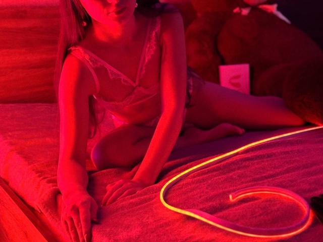 Rouzi vestindo lingerie vermelha, sentada em uma cama com uma luz de coração neon no cobertor, com os seios à mostra, iluminação vermelha, um brinquedo de pelúcia marrom ao fundo..