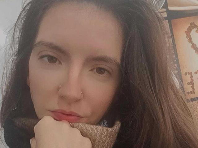 BraniaRuben has a nose piercing, brown eyes, and dark hair. She's holding a brown scarf with her hand, looking serious. Her lips are slightly red.