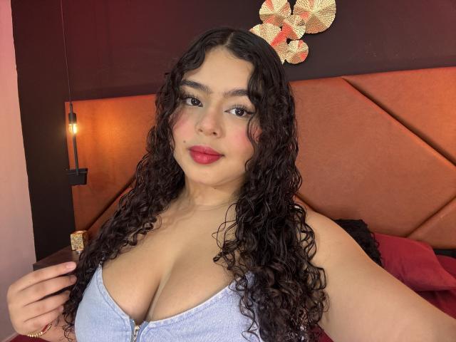 AprielKeith has curly black hair, red lipstick, and is wearing a blue zip-up top, showing her big tits. She's smiling at the camera, with a warm light behind.
