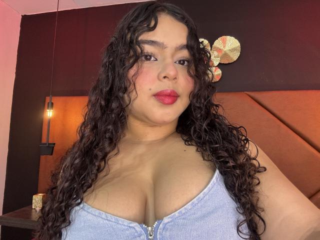 AprielKeith with curly black hair, pink lips, and blue denim top, showing her big tits and light brown skin, standing in a room with warm lighting.