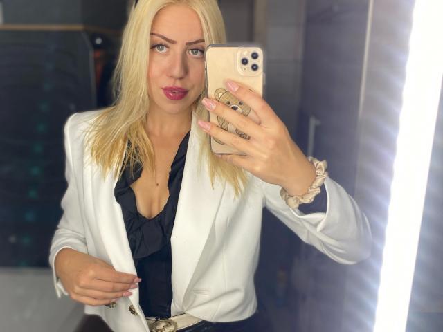 CarolyneMarianX-hot with blonde hair, white blazer, black ruffled shirt, pink lipstick, holding iPhone, scrunchie on wrist, taking mirror selfie, visible boobs, attractive, stylish.