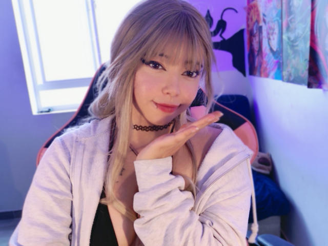 Arieyaara with blonde hair, bangs, and eyeliner, wearing a white zip-up, black choker, and black top, hand raised, smiling, in a brightly lit room.
