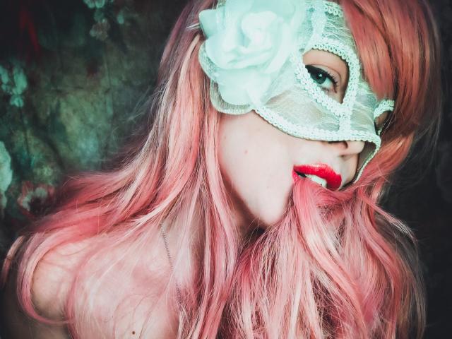 JuliaFrancaise, com cabelo rosa, máscara de renda branca e batom vermelho, com os seios quase imperceptíveis, olhando sedutivamente para a câmera. Ela tem uma rosa na máscara..