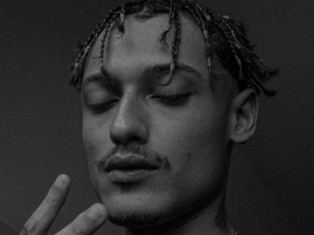 Black-and-white photo of BerninBoss with eyes closed, small mustache, and braided hair, hand gently touching chin. BerninBoss has a calm, focused expression.