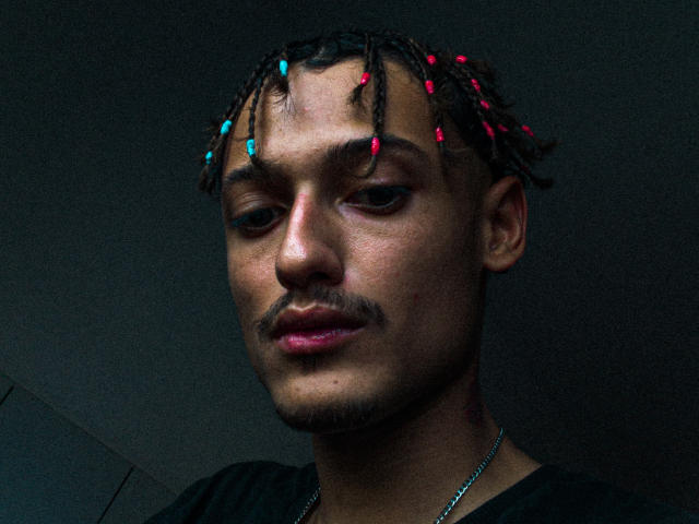 BerninBoss has dark skin, medium-length dreadlocks with red and blue beads, a mustache, and a chain necklace, looking down with a slightly serious expression.