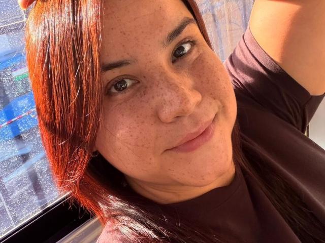 GabyTits has red hair, freckles, and brown eyes, wearing a maroon shirt, smiling slightly with sunlight on her face, raising her arm in a selfie.