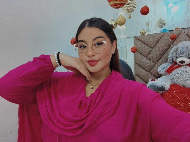HelueJamil with glasses, pink blouse, and red lipstick, resting her chin on her hand, smiling. She has a cute, sexy look, dark hair, and a small gold necklace.