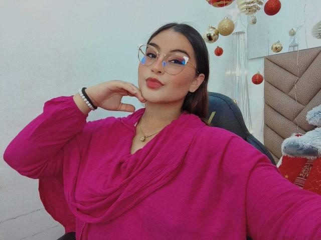 HelueJamil with dark hair, glasses, pink shirt, and necklace, holding cheek, smiling, showing off her tits, sitting in a chair with a red bag.