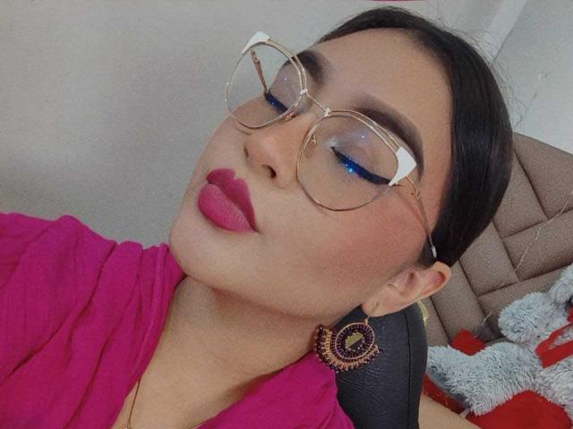 HelueJamil with puffy pink lips, blue eyeshadow, gold-rimmed glasses, and pink top, wearing circular earrings, head tilted back, black chair in background, dark hair tied back.