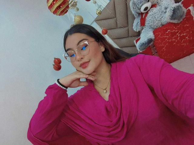 HelueJamil with glasses, pink cowl-neck top, red lips, hand on chin, wearing bracelet and necklace, next to stuffed gray cat on red pillow, Christmas decorations in background.