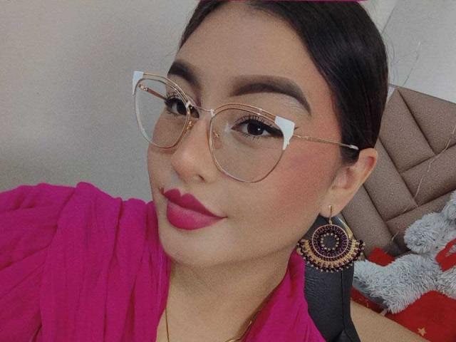HelueJamil has dark hair, pink lipstick, and glasses with white accents. She's wearing a pink top and large, decorative earrings. Her makeup is bold, and she's smiling.