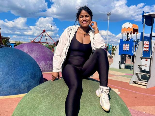 IvannaDuran with dark tits and ass in black leggings, white jacket, and white sneakers, sitting on green play structure, smiling, under blue sky with clouds.