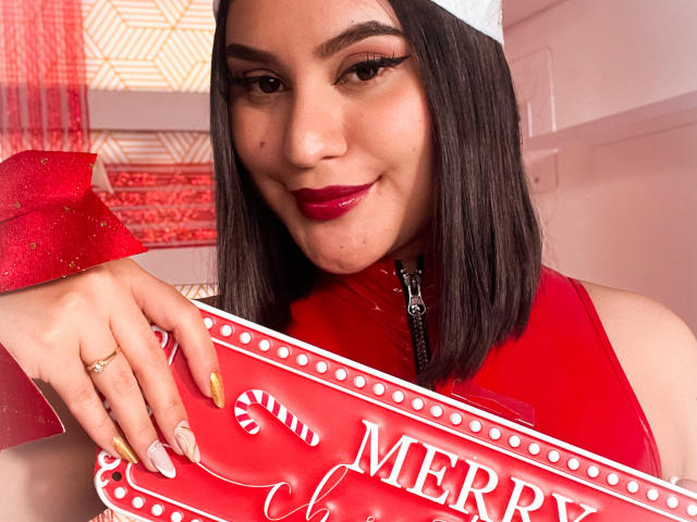 AdaKolex with dark brown bob, white headband, red lipstick, and gold nails, holding a red candy cane tray, smiling, wearing a red sleeveless top, showing a ring on her left hand.