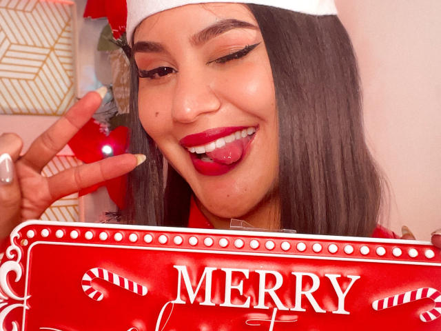 AdaKolex with long black hair, red lipstick, and white hat, tongue out, winking, holding red candy cane sign, making peace sign, has cute nose mole, and painted nails.