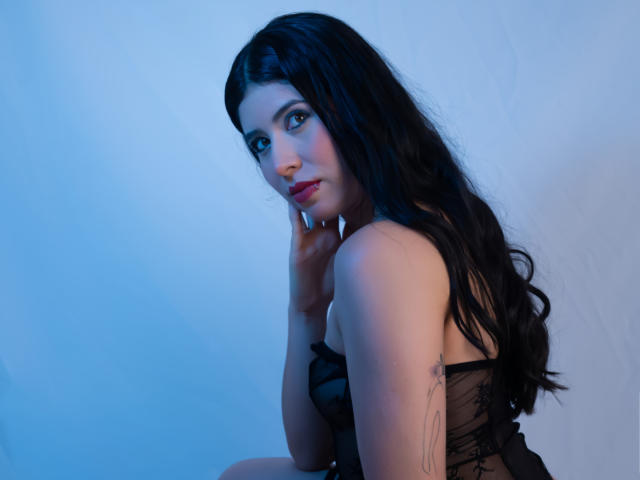 MiluSky with long black hair, blue eyes, and red lipstick, wearing black lingerie, has her hand on her cheek, showing a tattoo on her right arm, in a blue-lit shot.