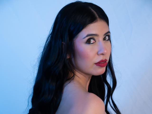 MiluSky with long black wavy hair, red lipstick, and subtle makeup, looking over her shoulder with a slight smile. She has a light skin tone and is topless.