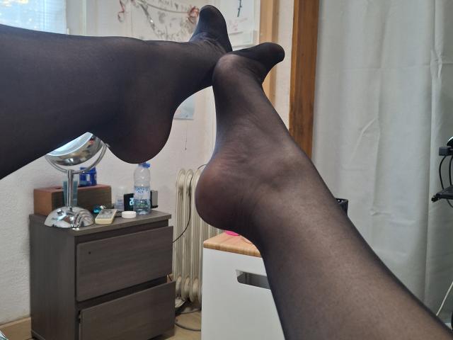 LaFrancaiseJessy's legs, wearing sheer black pantyhose, crossed at the ankles, showing her soles and toes, with a water bottle and small mirror on a wooden dresser.