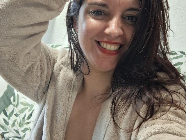 LaFrancaiseJessy with wet brown hair, smiling with red lipstick, wearing a beige fluffy robe, showing her white teeth, and raising her right arm. Her tits are slightly visible.