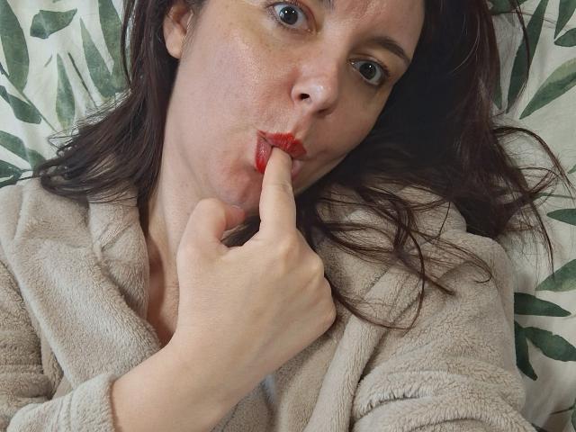 LaFrancaiseJessy with red lipstick, puffy beige robe, and finger in mouth, brown hair, green eyes, slightly freckled face, lying down, looking at camera.