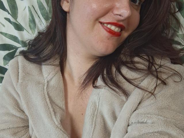 LaFrancaiseJessy has red lipstick, a cute smile, and is wearing a fluffy beige robe. Her brown hair is loose, and she has fair skin.