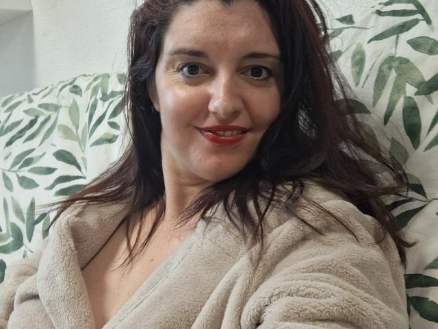 LaFrancaiseJessy with red lipstick, medium tits, and brown wavy hair, smiling in a beige fluffy robe, lying on a green leaf patterned pillow.