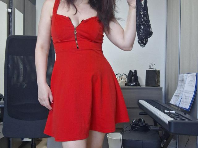 LaFrancaiseJessy in a red dress with a zipper, holding black lace underwear, standing in a room with a keyboard and chair, showing her tits and ass.