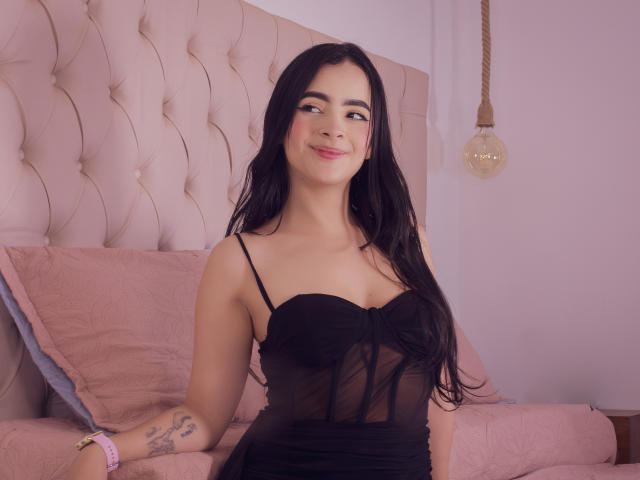 KonanStar with long black hair, wearing a sheer black dress, sitting on a pink bed, smiling, showing small tattoos on her right arm, and a light bulb in the background.