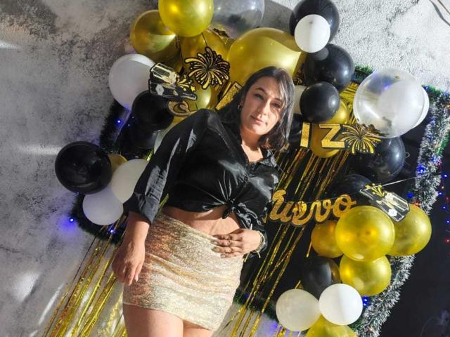 EmaFoxy with small tits, wearing a black shirt tied at the front and a glittery gold skirt, standing in front of a bunch of black, white, and gold balloons.