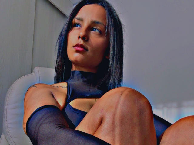 Loren, avec de longs cheveux noirs, des yeux bleus et du rouge à lèvres rose, portant un haut noir transparent et des manchettes, assise les genoux repliés, regardant sur le côté..