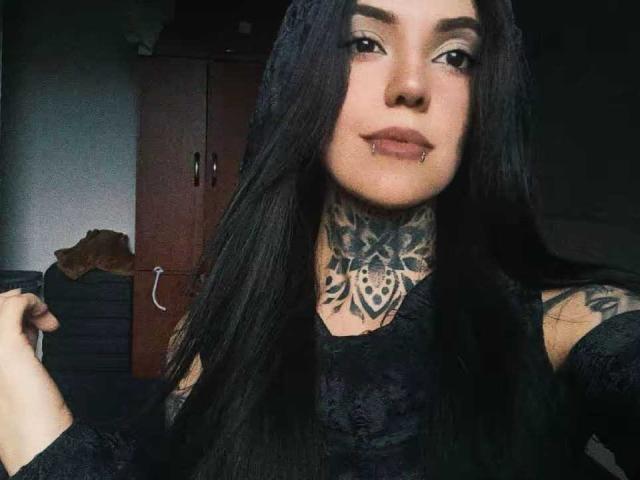 Lannie Gonzales, com cabelos pretos longos, tatuagens no pescoço e no peito, blusa de renda preta, um leve sorriso, um piercing no nariz e segurando o cabelo. Quarto escuro, armário de madeira ao fundo..