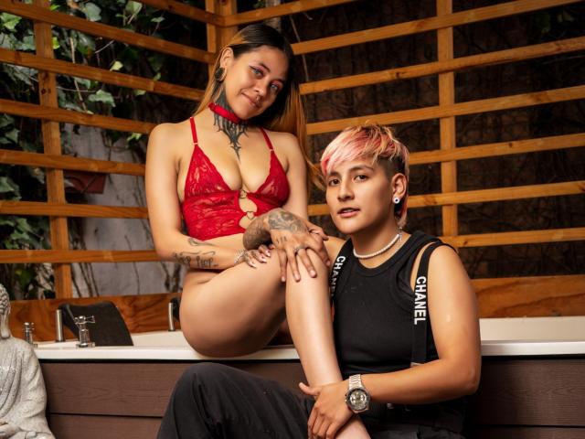 HadeDanny with red lace bra and tits, sitting on a counter, tattooed arm on another HadeDanny's leg, short pink hair, black tank top, wooden wall, relaxed expression.