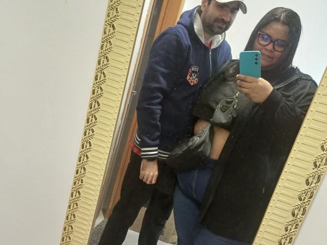 ReeseDuo met bril, zwart haar, zwarte jas, blauwe jeans, met een zwarte handtas, staat naast een man met baard in een blauwe hoodie, pet, zwarte broek, en maakt een selfie in de spiegel..