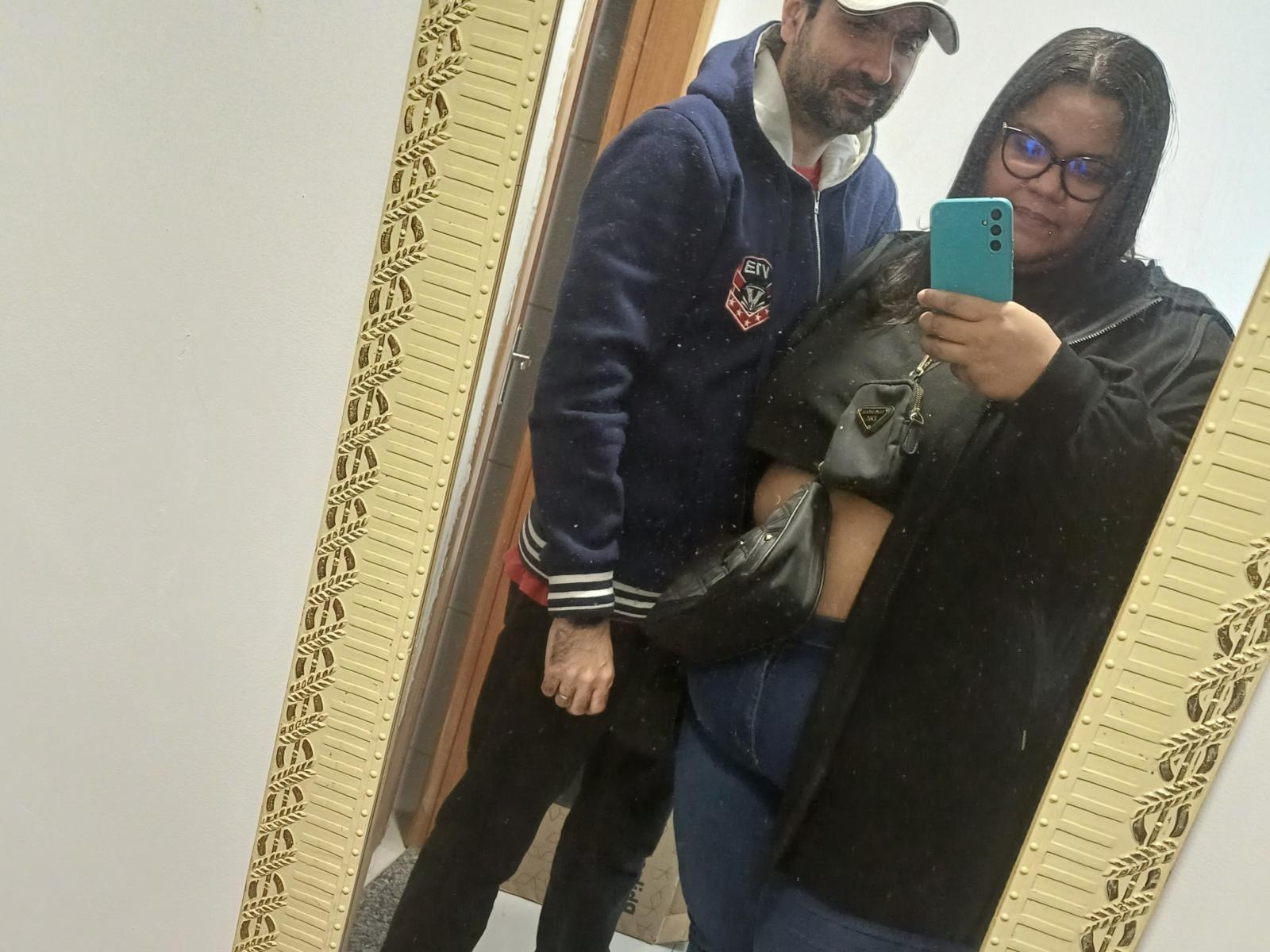 ReeseDuo with glasses, black hair, black jacket, blue jeans, holding black purse, standing next to bearded man in blue hoodie, cap, black pants, taking mirror selfie.