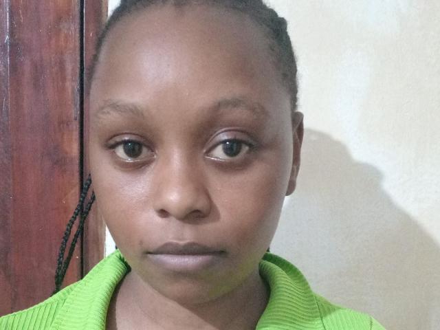 MadonahLynn has dark brown skin, small round eyes, and a serious expression. She's wearing a green ribbed shirt, with her braids pulled back. She's staring at the camera.