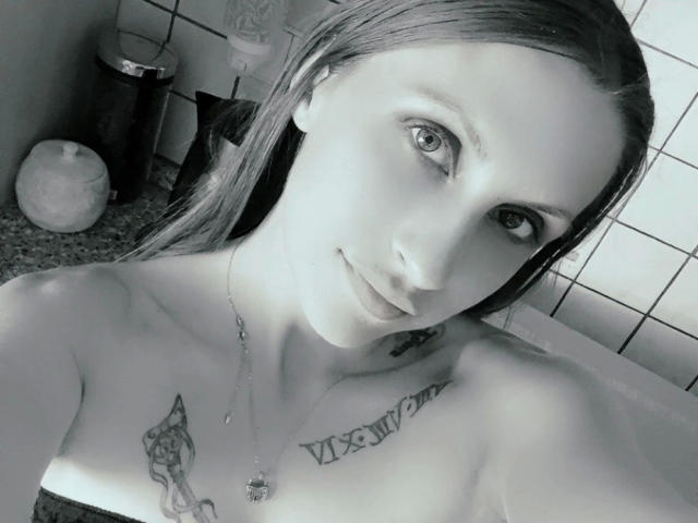 Black-and-white selfie of Joie-20-hot with wet hair, looking at camera, showing tattoos on chest and collarbone, necklace, and bare shoulders, with a slight smile.