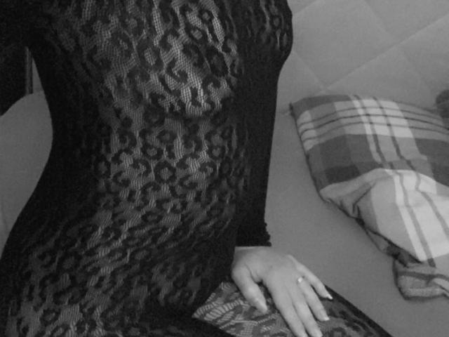 Joie-20-hot in a black lace dress, showing her tits through the patterned fabric, sitting with a hand on her hip, a plaid pillow in the background.