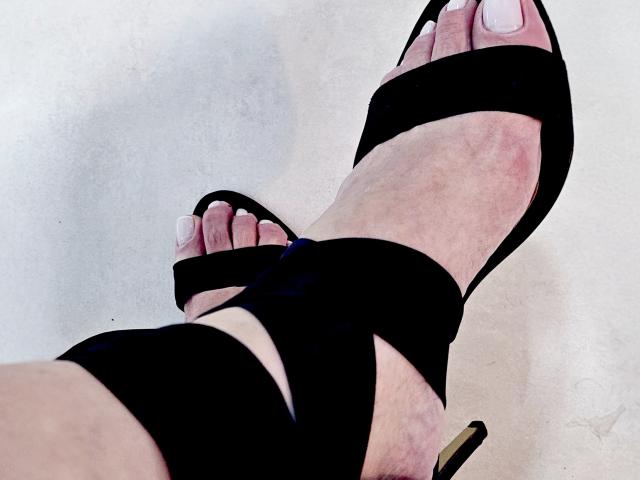 Les pieds de CherryBores sont chaussés de talons hauts noirs à brides, avec des ongles peints en blanc, et le talon est doré en bas à droite. Sa peau est claire, et l'attention est portée sur ses pieds..