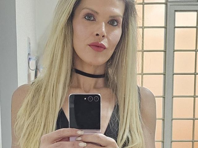 CherryBores met blond haar, die een zwarte choker en een top van kant draagt, een telefoon vasthoudt, de camera aankijkt met roze lippenstift, en haar borsten laat zien..
